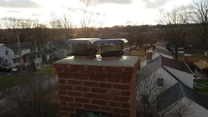 Brick chimney with two metal chimney caps against a neighborhood backdrop at dusk.