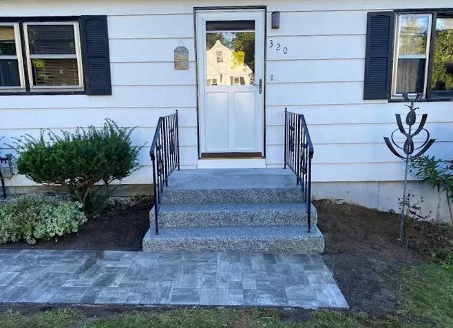 White house with gray steps leading to a white door. Black handrails, brick patio, and landscaping.