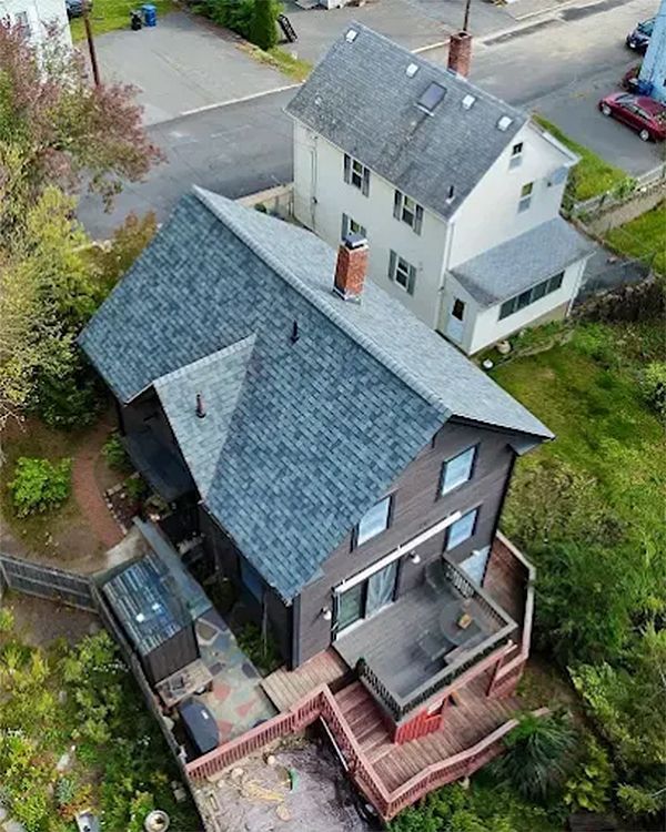 Aerial view of two-story brown house with deck and gray roof; adjacent beige house with gray roof.