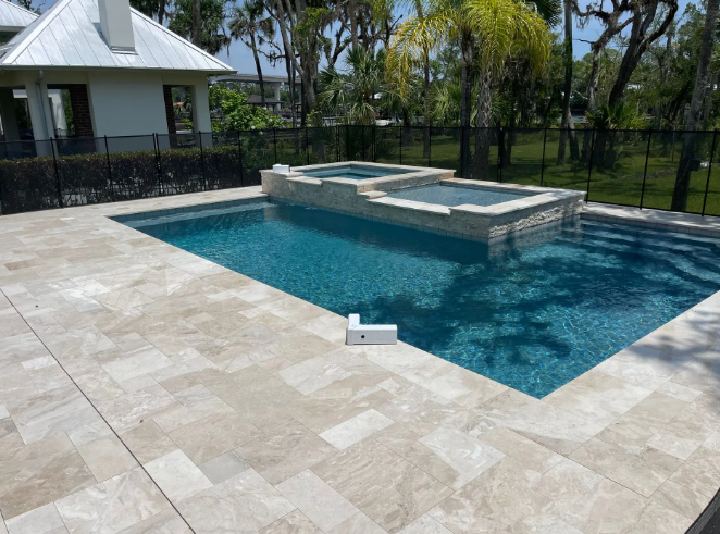 Pool with attached spa, surrounded by stone patio, black fence, and white house.