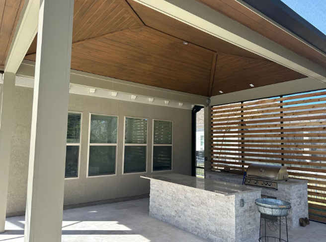 Outdoor kitchen with a bar, grill, and wooden slatted wall. Brown ceiling, gray countertop.