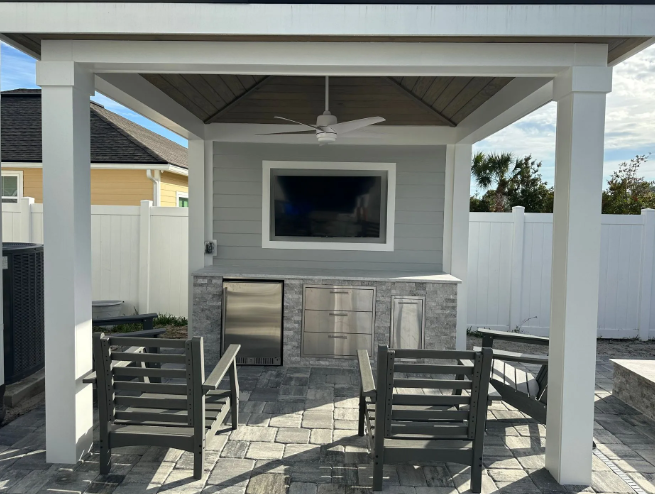 Outdoor kitchen with gray cabinets, large TV, fan, and seating under a white pergola.