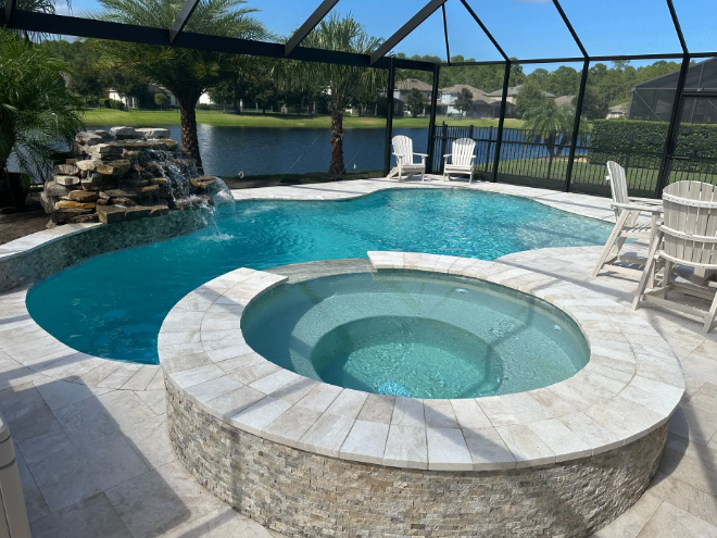 Pool with waterfall, spa, and seating in a screened-in patio overlooking a lake.
