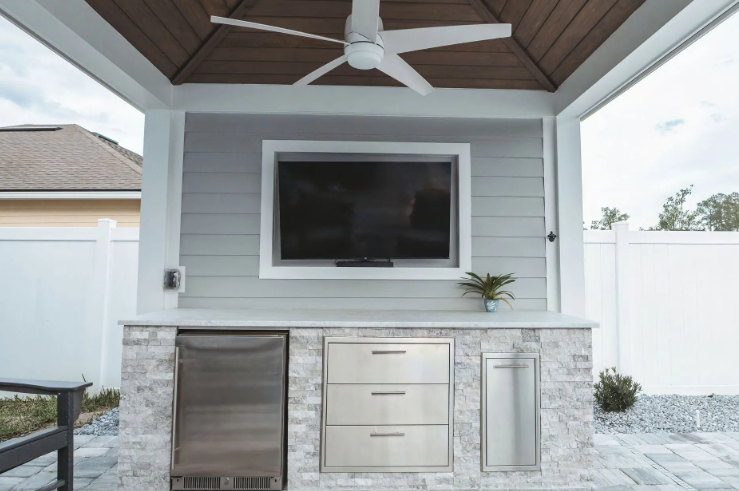 Outdoor kitchen with TV, stainless steel appliances, and ceiling fan under a covered patio.
