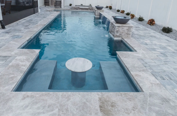 Long, rectangular pool with submerged seating and table; surrounded by gray tiled patio.