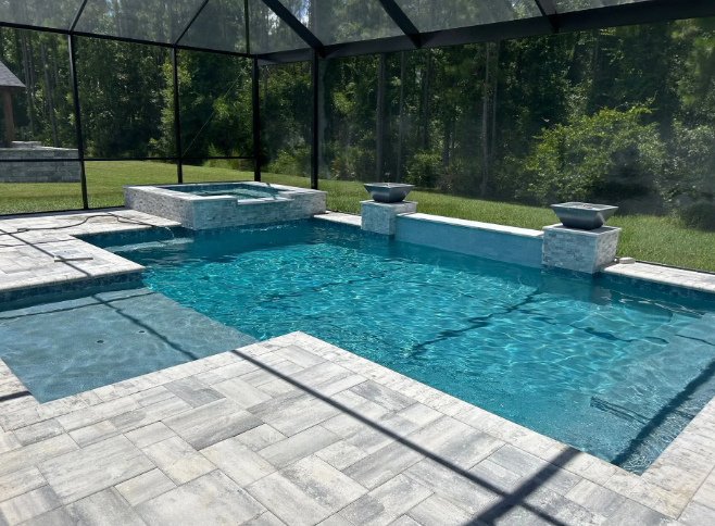 A pool with turquoise water, a hot tub, and paver patio enclosed by a screen. Green grass and trees in the background.