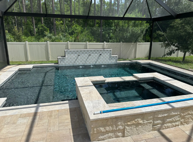 Pool and hot tub enclosed in a screened-in structure with a waterfall feature. Beige stone and dark water.