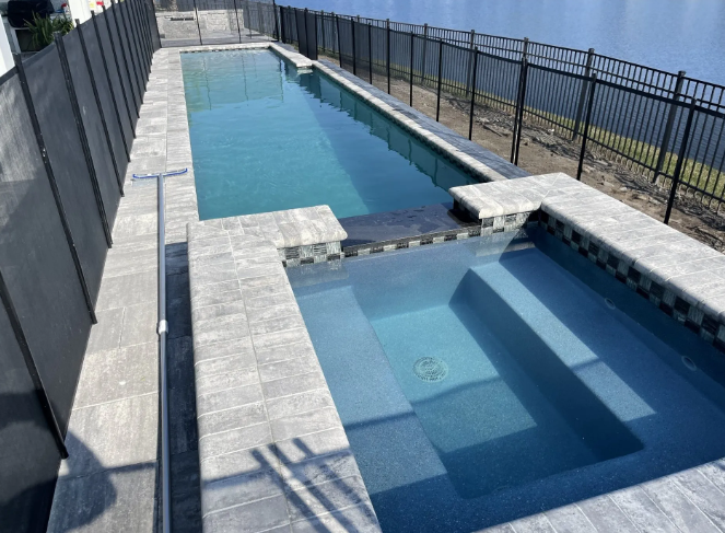Pool with rectangular design, featuring a long swimming area and a square spa, surrounded by grey stone and black fence.