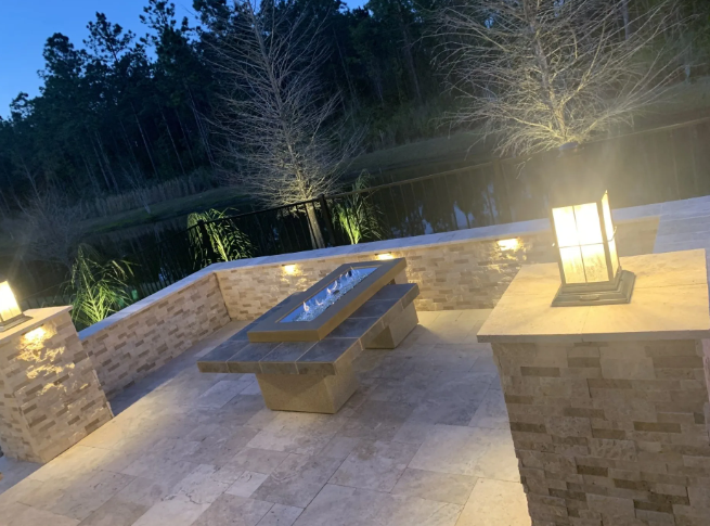 Outdoor patio with a fire pit, illuminated by ambient lighting, overlooking a dark lake.