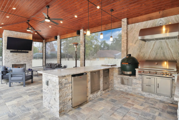 Outdoor kitchen with stone counters, a grill, and a fireplace under a wood-paneled ceiling.