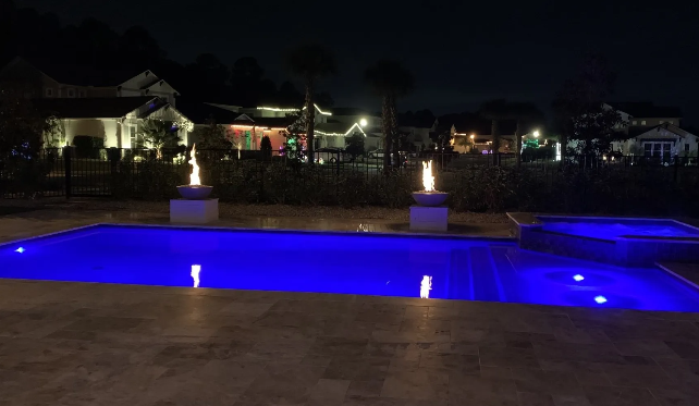 Nighttime pool scene: blue-lit pool, two fire features, homes in the background.