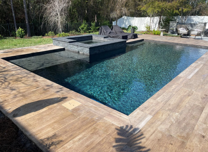 Outdoor pool with dark blue water and light brown stone edging; lounge chairs are visible in the background.