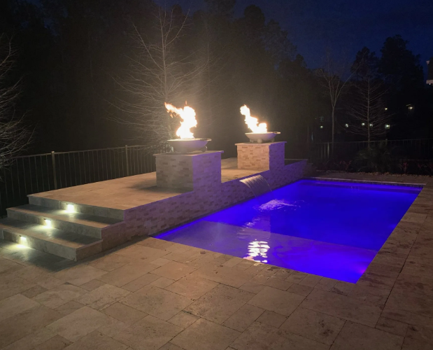 Nighttime backyard pool with lit fire bowls, steps, and blue underwater pool lighting.