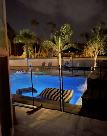 Nighttime backyard pool scene with illuminated palm trees, floating rafts, and chairs.