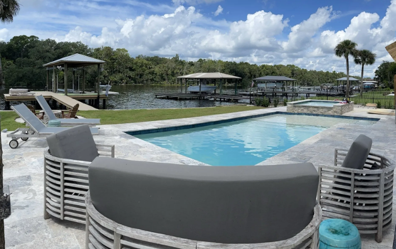 Poolside view with pool, lake, and docks. Gray outdoor furniture. Sunny day.