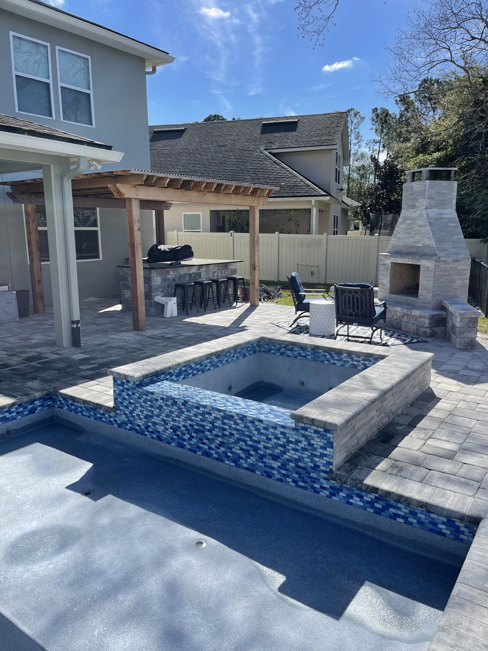 Backyard with pool, hot tub, outdoor kitchen under pergola, and stone fireplace on a sunny day.