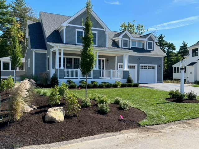 Gray two-story suburban house with white trim, porch, garage, and landscaped front yard on a sunny day