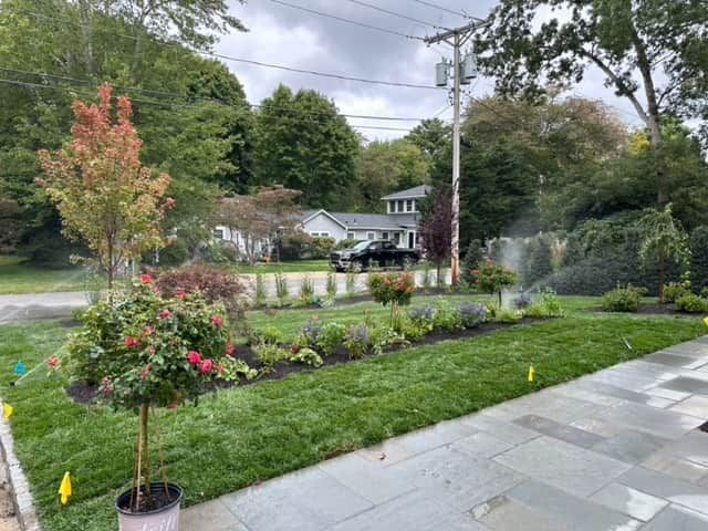 Landscaped garden with flowers, shrubs, lawn, and stone patio beside a road and trees