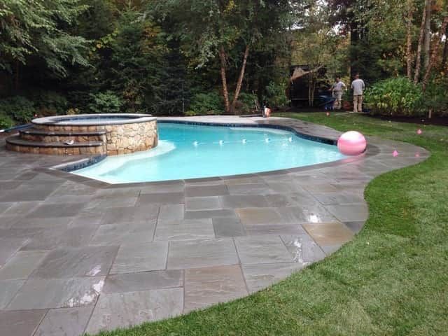 Backyard swimming pool with stone patio, hot tub, and pink float in a wooded yard