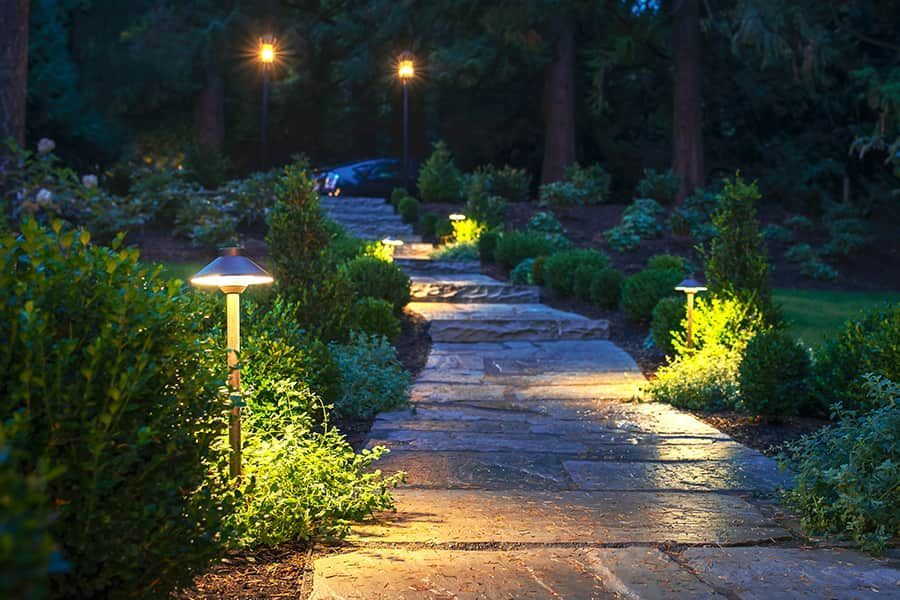 Lit garden path winding through lush greenery at dusk, with glowing pathway lights and a distant streetlamp