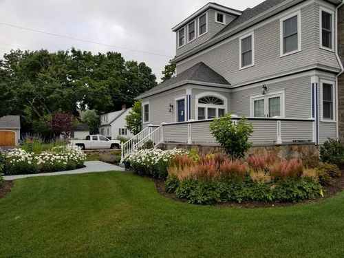 Gray house with white trim and manicured lawn, garden beds, and a walkway under cloudy skies