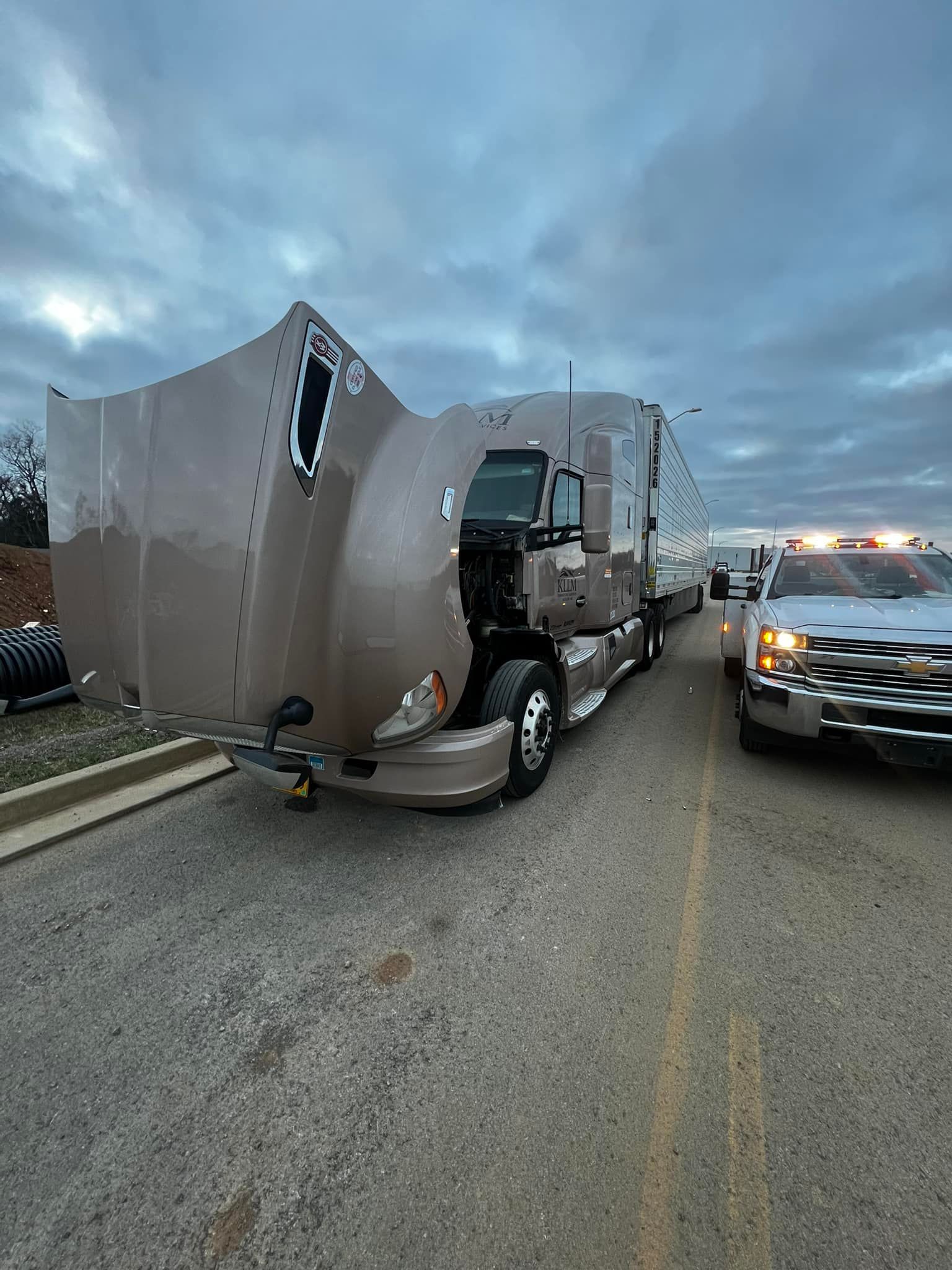 Damaged semi-truck on the side of the road, with a tow truck behind it, under a cloudy sky.
