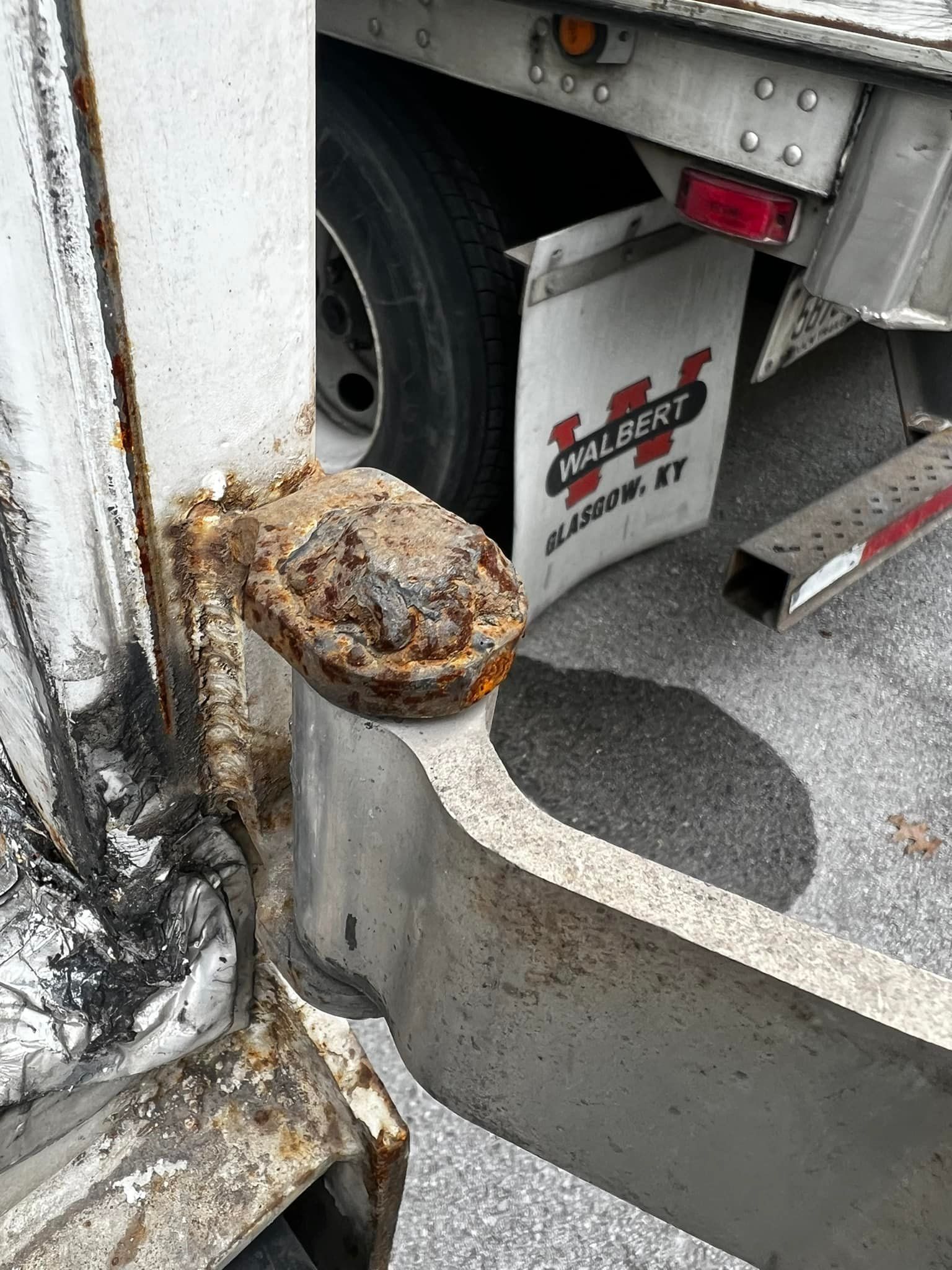 Rust-covered trailer hitch. Silver metal with a Wilbert Louisville, KY mud flap, a tire, and water stain.