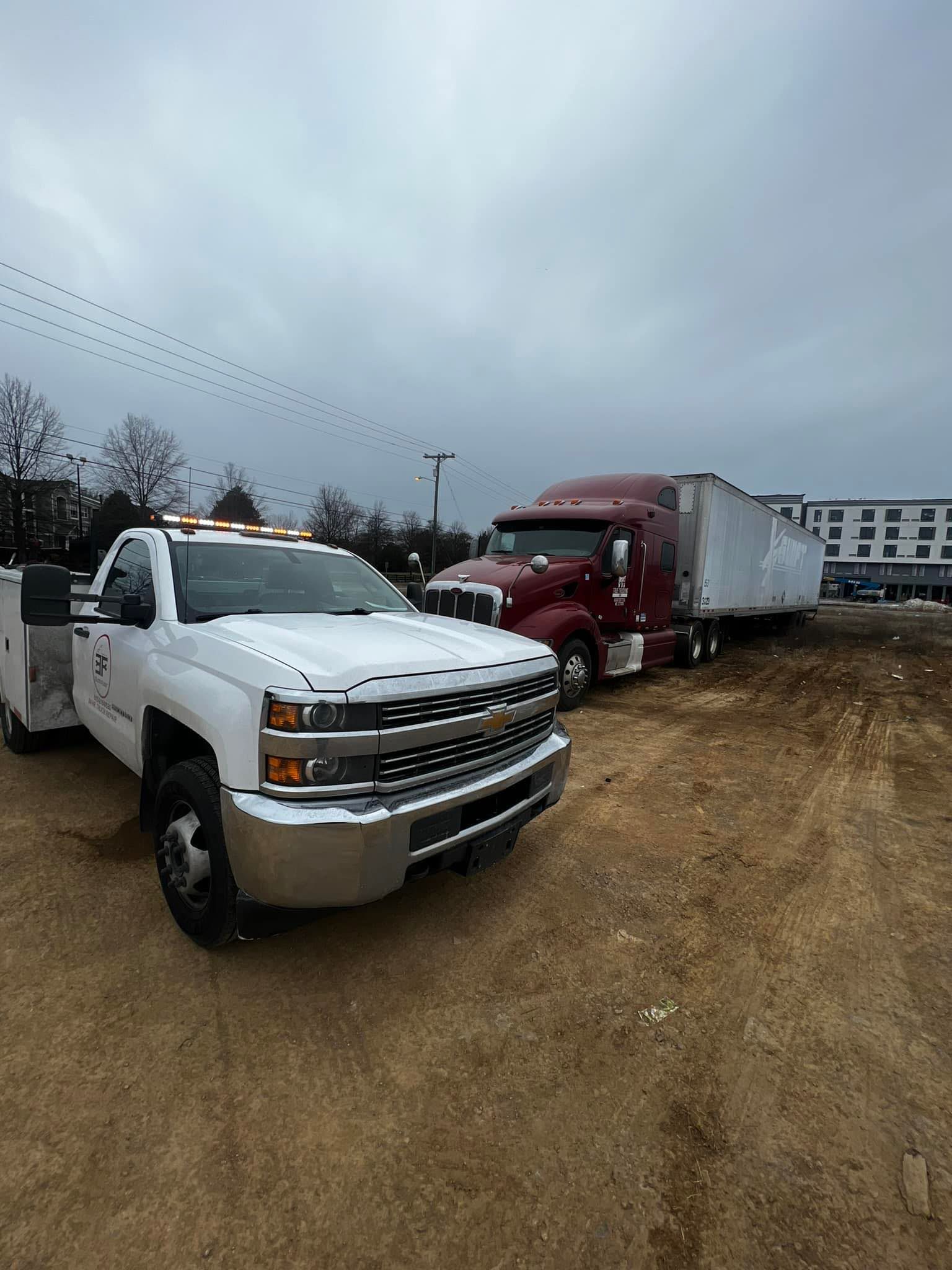 White tow truck towing a maroon semi-truck on a dirt lot under a cloudy sky.