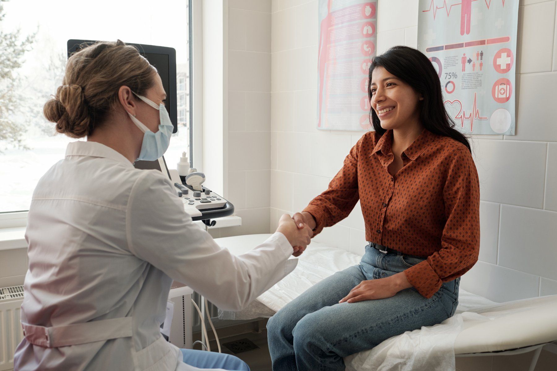 Doctor wearing a mask shakes hands with a smiling patient in an examination room.