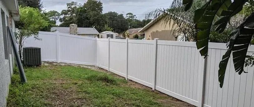 White vinyl fence in a backyard. An air conditioner and foliage are in view.