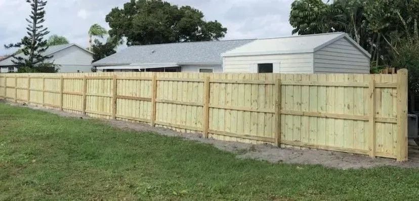 Wooden fence along a grassy yard, with houses in the background under a cloudy sky.