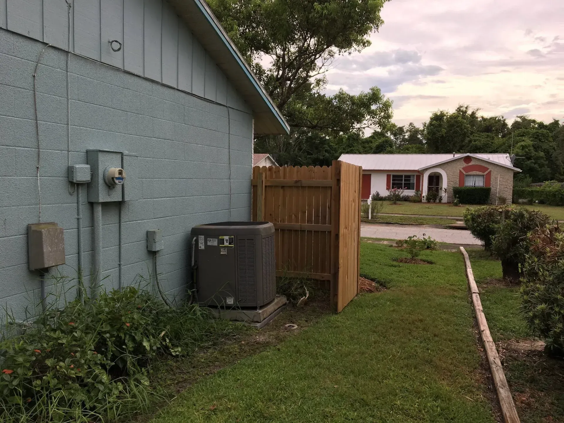 Exterior of a building with an AC unit and a small wooden fence; a house is in the background.