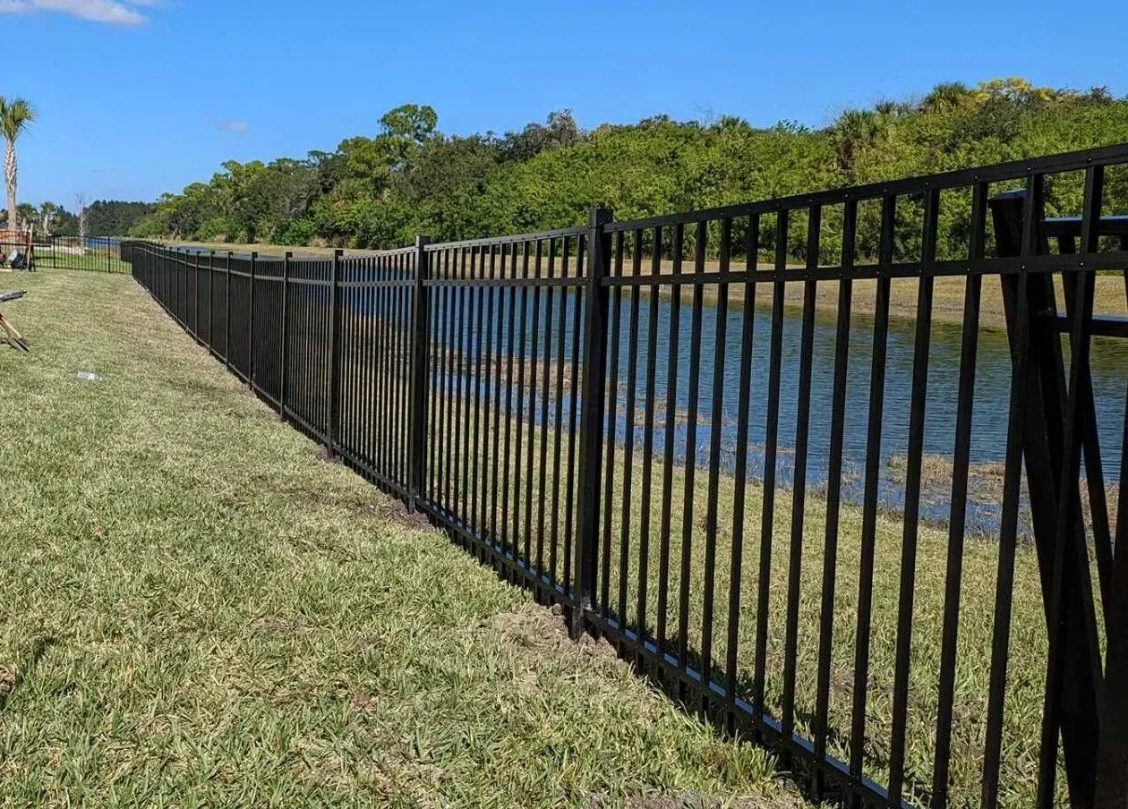 Black metal fence along a waterway, green grass, trees in the background, clear sky.