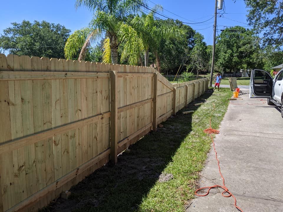 Wooden fence along a sidewalk, with greenery and trees in the background under a bright blue sky.