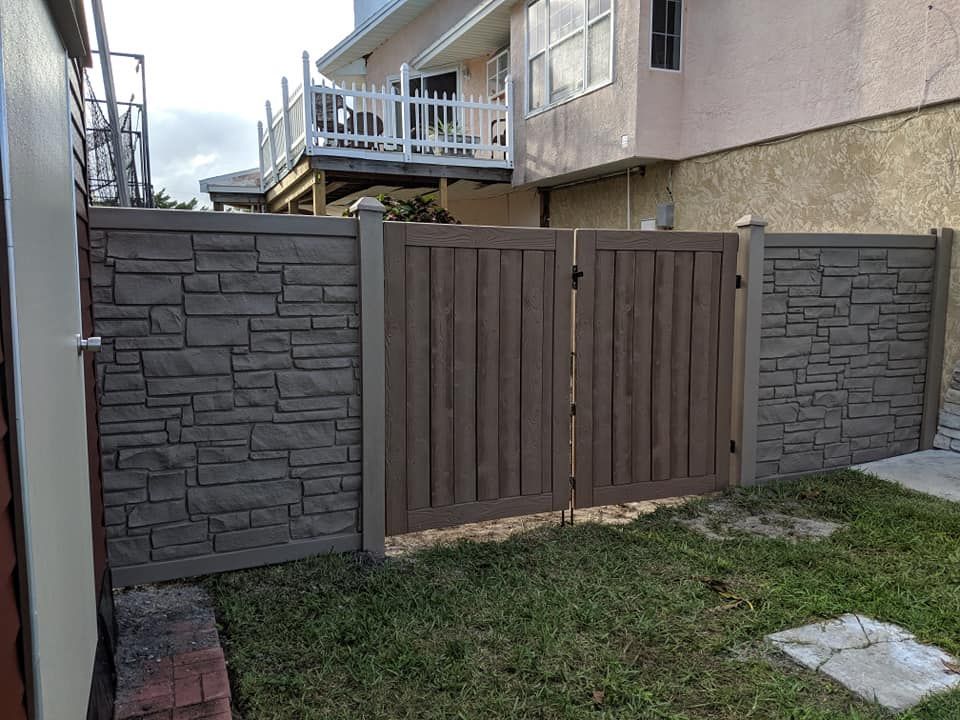 Brown wooden gate and stone-look fence enclosing a yard, next to a beige building.