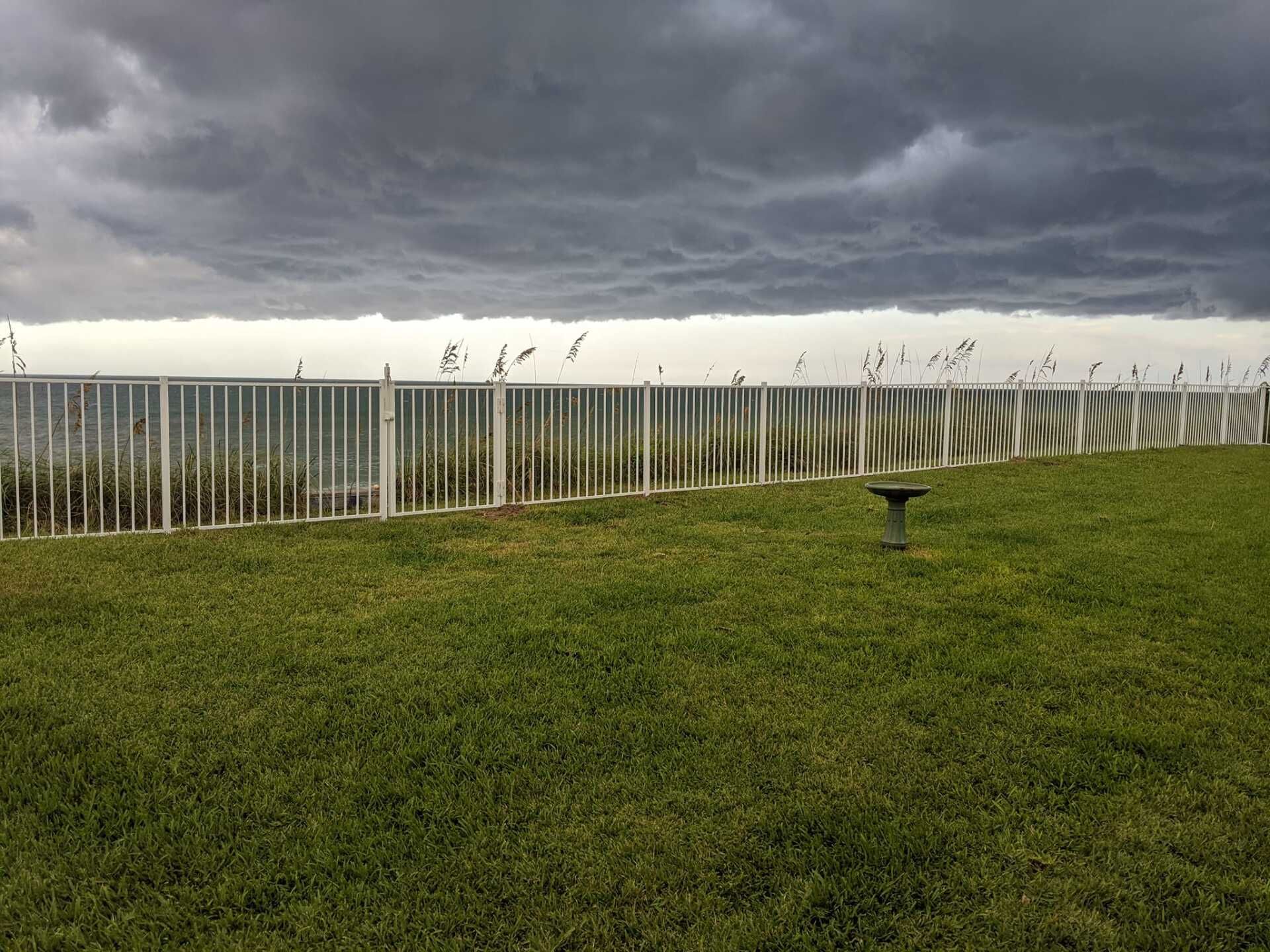 Lush green lawn with white fence, silhouetted grass, and ominous dark storm clouds overhead.