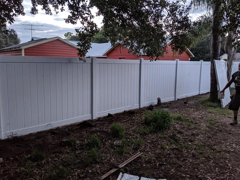 White vinyl fence in a backyard setting, with red houses visible in the background. A person stands nearby.