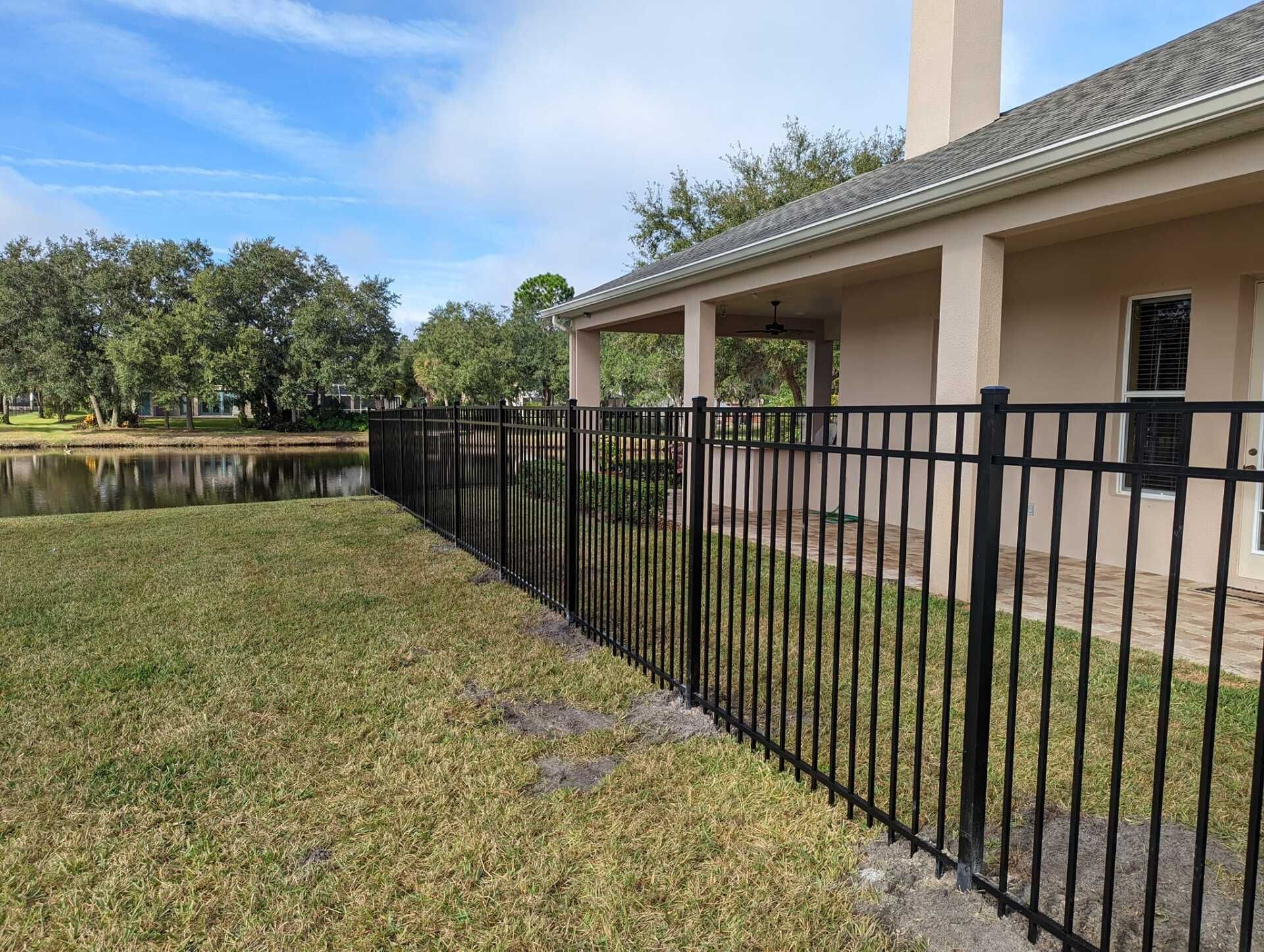 Black metal fence along a building, next to a grassy area and a body of water under a blue sky.