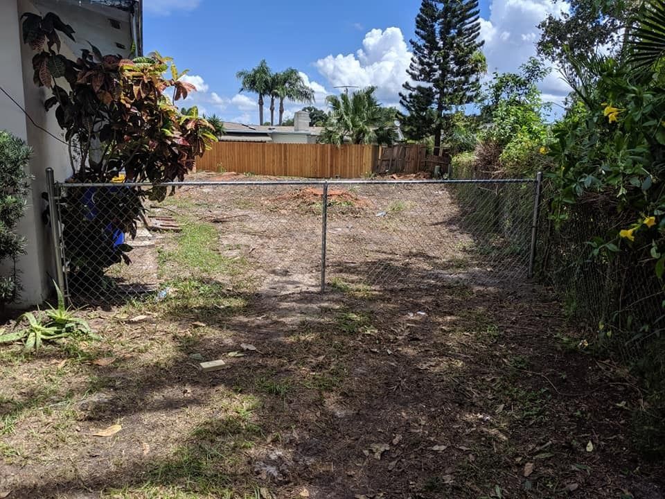 Chain-link fence encloses a bare dirt backyard. Wooden fence and trees in background. Cloudy sky.