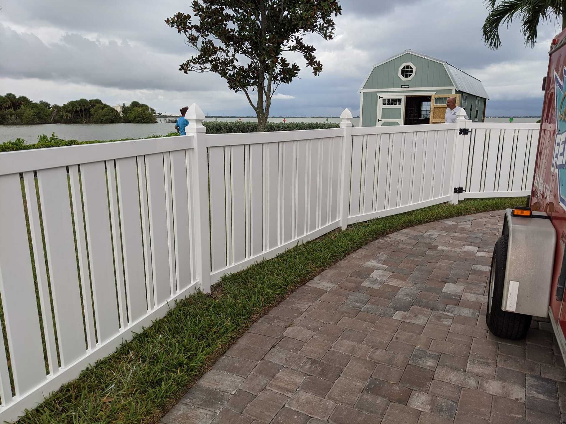White fence curving along a brick path, with a green shed and water in the background.