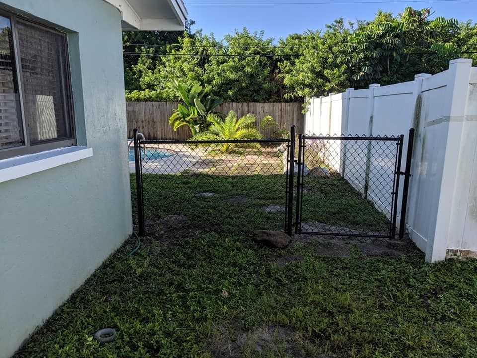 Backyard with chain-link fence, gate, and white vinyl fence; grass and greenery surround.