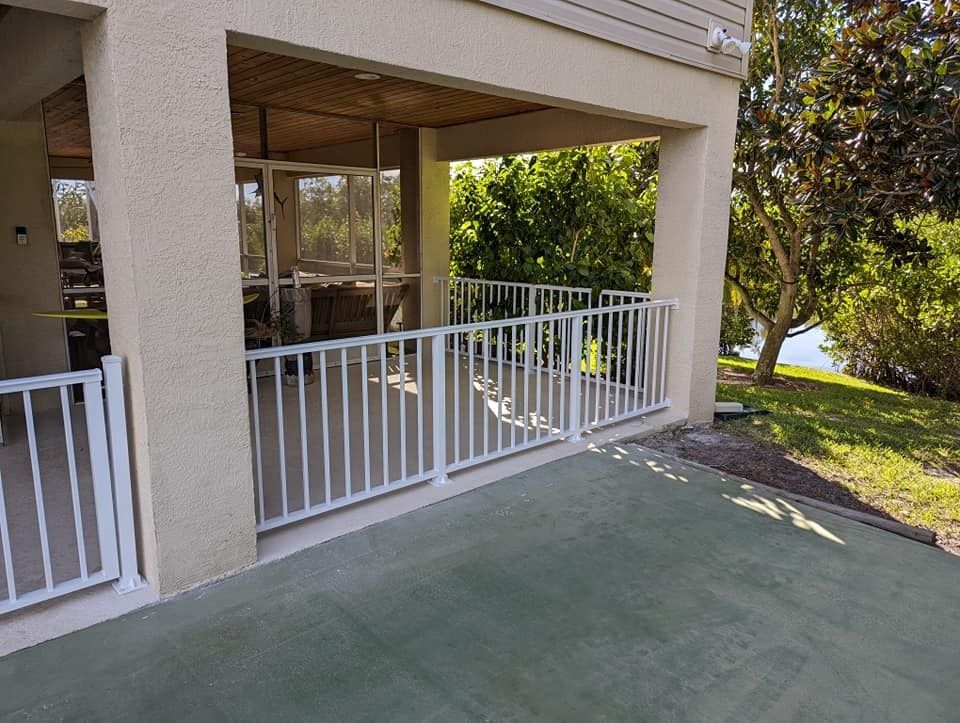 White railing encloses a patio under a building. Green patio floor, view of water and trees.