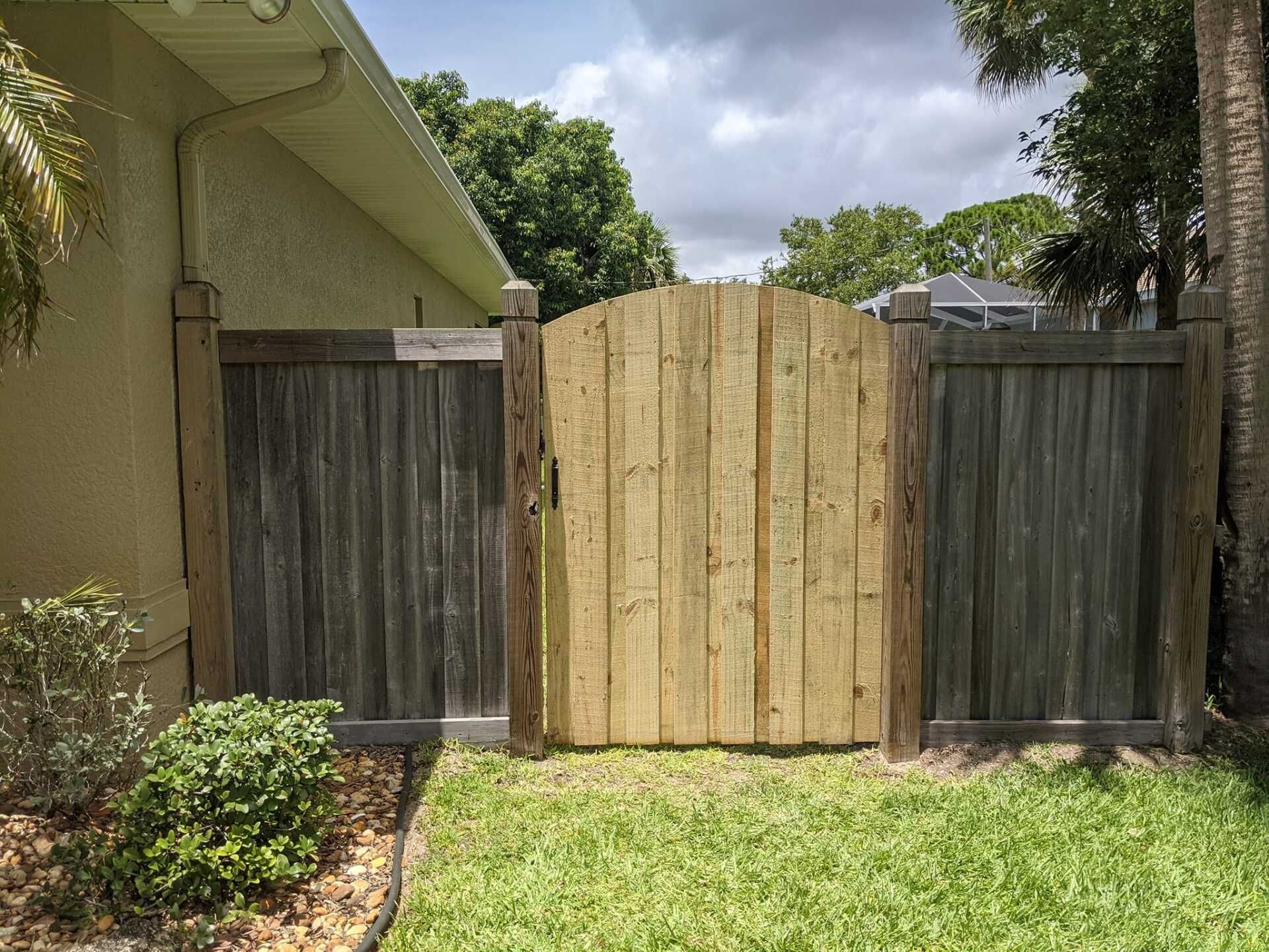 Wooden gate in a backyard between two weathered fences, with green grass and foliage.