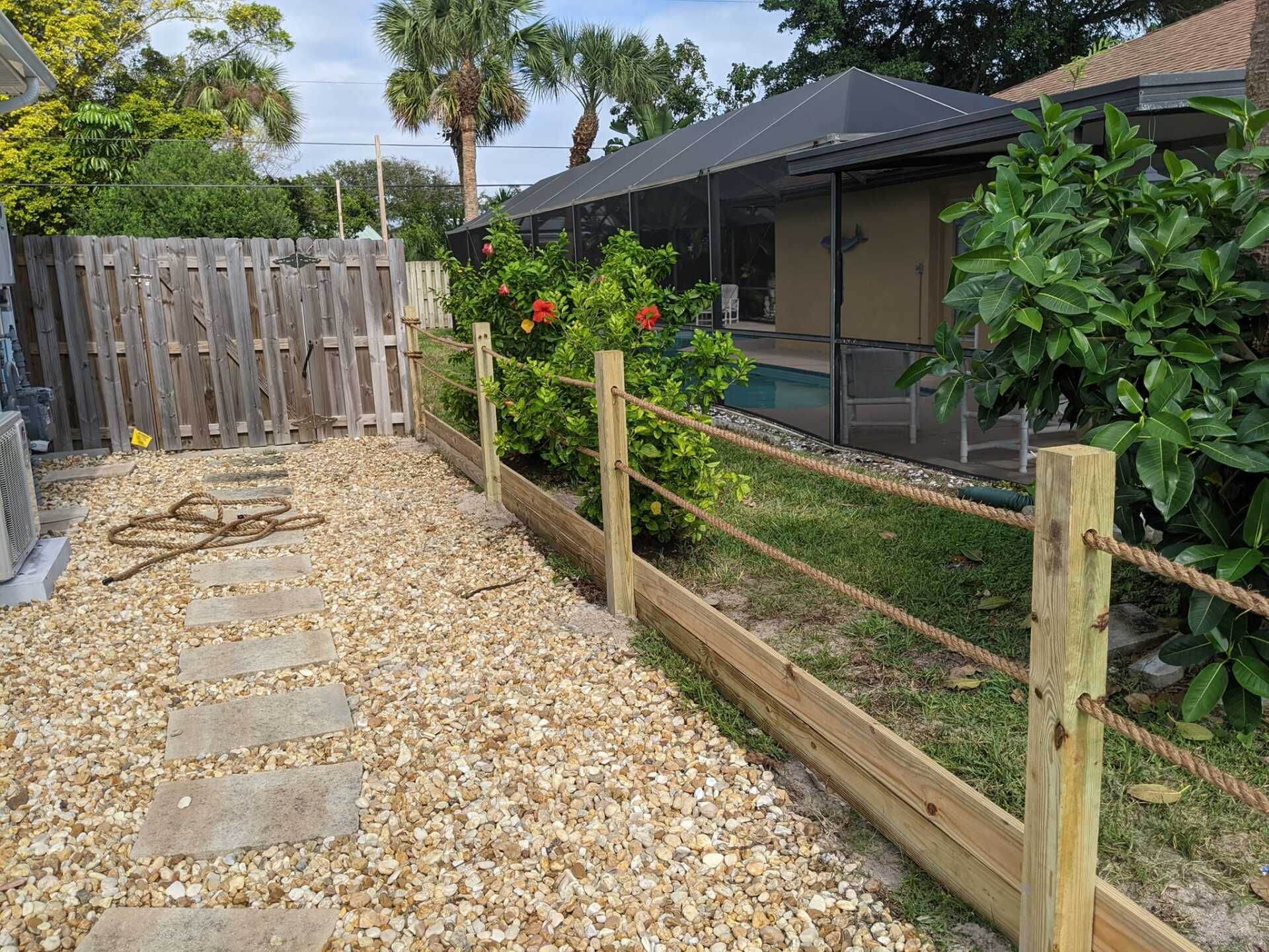 Backyard with stone path, wood fence, and rope border near a screened-in pool. Green plants and trees.