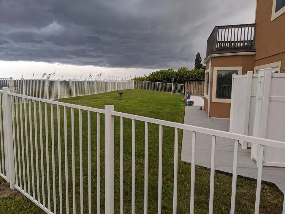 White fence surrounds a green lawn next to a two-story house, under a stormy gray sky.
