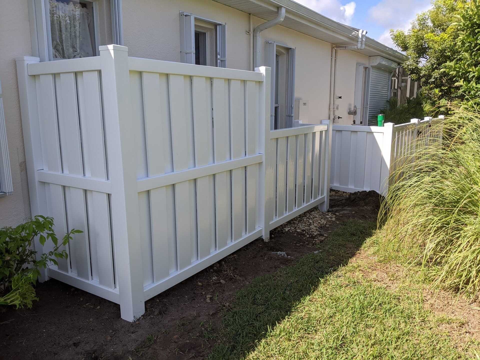 White vinyl fence outside a light-colored building, with green grass and foliage nearby.