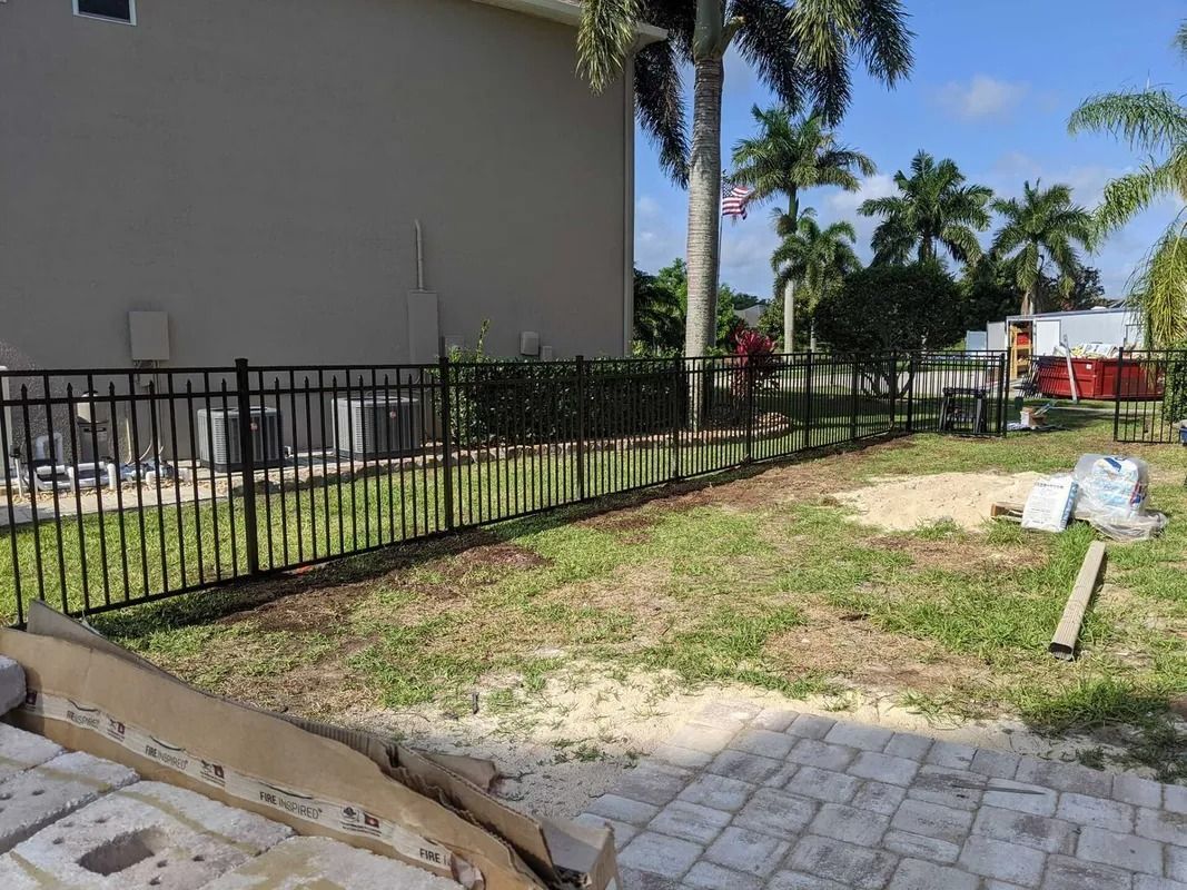 Black metal fence bordering a yard next to a beige building, with a red dumpster in the background.