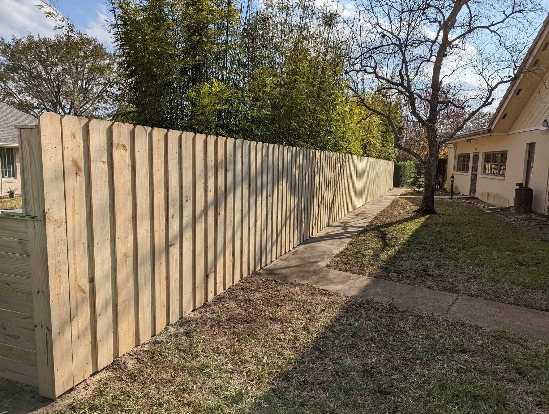 Wooden fence in a yard, beside a concrete walkway, with trees and a house in the background.