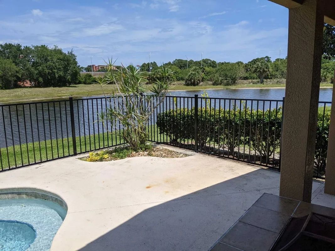 Patio with a pool overlooking a lake, surrounded by a black fence and greenery.