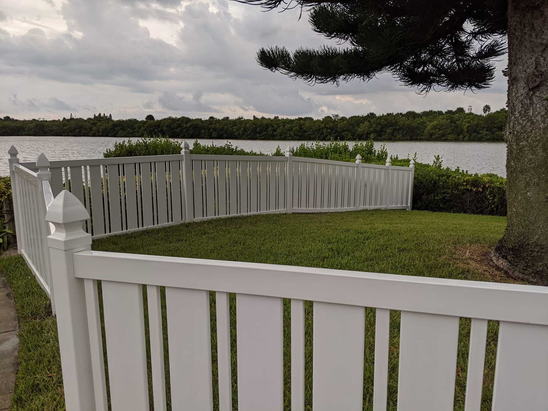 White fence borders a grassy area near a lake. Trees and cloudy sky in background.